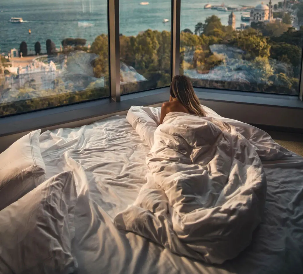 A woman laying in bed with sheets on and looking out of the window.